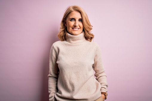 Middle age beautiful blonde woman wearing casual turtleneck sweater over pink background with a happy and cool smile on face. Lucky person.