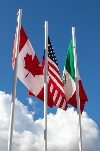 Flags of 3 USMCA countries consisting of United States, Mexico, Canada on flag pole against sky, concept of new NAFTA agreement that is known as USMCA in the U.S., CUSMA in Canada or T-MEC in Mexico.