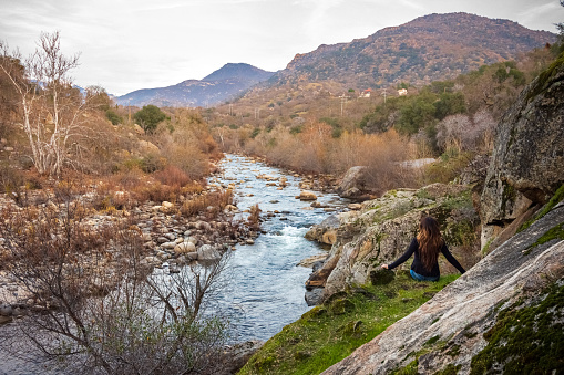 A relaxed woman is enjoying the view of Sierra Nevada mountains from the banks of the Kaweah River in Three Rivers, CA. The river begins as four forks in Sequoia National Park, home to the largest trees on Earth. It then flows in a southwest direction to Lake Kaweah. Shot in Winter.