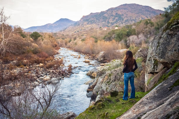 A relaxed woman is enjoying the view of Sierra Nevada mountains from the banks of the Kaweah River in Three Rivers, CA. The river begins as four forks in Sequoia National Park, home to the largest trees on Earth. It then flows in a southwest direction to Lake Kaweah. Shot in Winter.