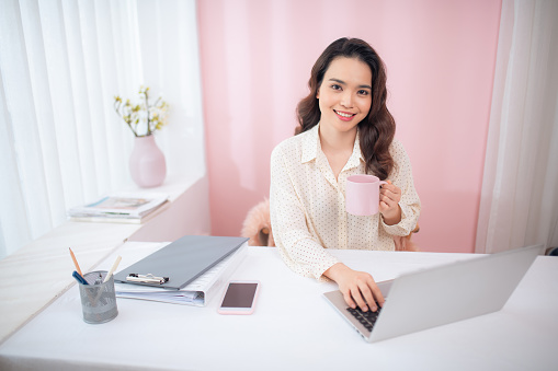 Beautiful Asian woman using laptop at her office while holding tea mug.