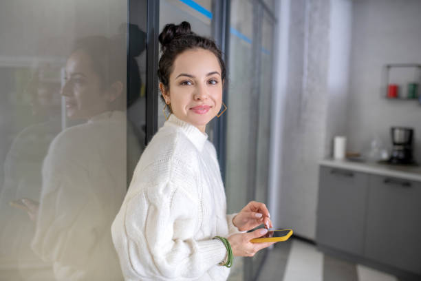 istock Young woman leaning on glass wall, holding mobile, smiling 1212310002