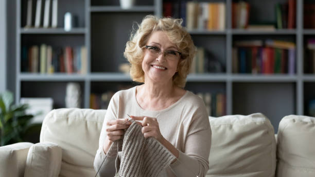 Smiling 60s grandmother enjoy knitting at home Portrait of smiling mature 60s grandmother sit on couch in living room knitting using needles, happy middle-aged 70s woman relax at home do favorite hobby activity on weekend, wellbeing concept grandmother stock pictures, royalty-free photos & images