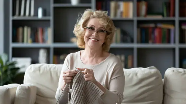 Portrait of smiling mature 60s grandmother sit on couch in living room knitting using needles, happy middle-aged 70s woman relax at home do favorite hobby activity on weekend, wellbeing concept