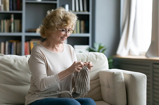 Happy middle-aged woman sit on sofa in living room hold needles knitting at home, relaxed elderly grandmother enjoy domestic weekend engaged in favorite hobby activity, retired wellness concept