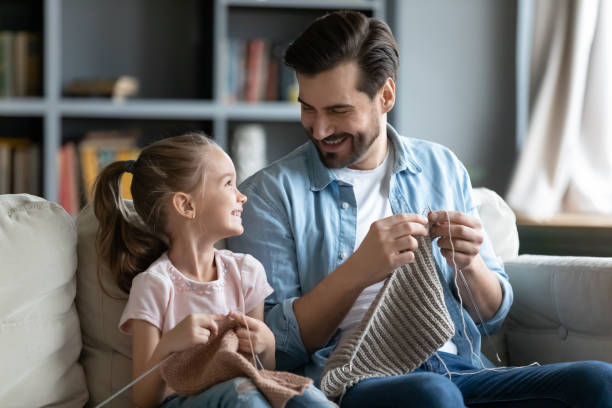 Smiling father and little daughter knitting at home together Smiling young father and little preschooler daughter sit on couch in living room knit with needles together, happy dad and small girl child have fun involved in favorite hobby family activity at home knitting photos stock pictures, royalty-free photos & images