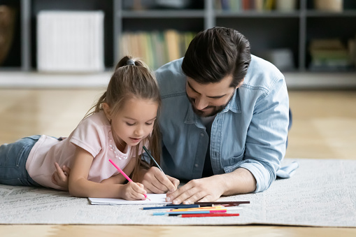 Young father lying on wooden floor with little preschooler daughter drawing together, caring dad have fun relax with small girl child paint in album, family hobby activity, early development concept