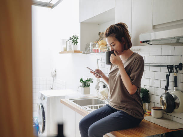 Young woman sitting in cozy kitchen and working on her mobile. Teenage asian woman drinking coffee while sitting on kitchen counter and working on smart phone in morning at home. coffee drink stock pictures, royalty-free photos & images
