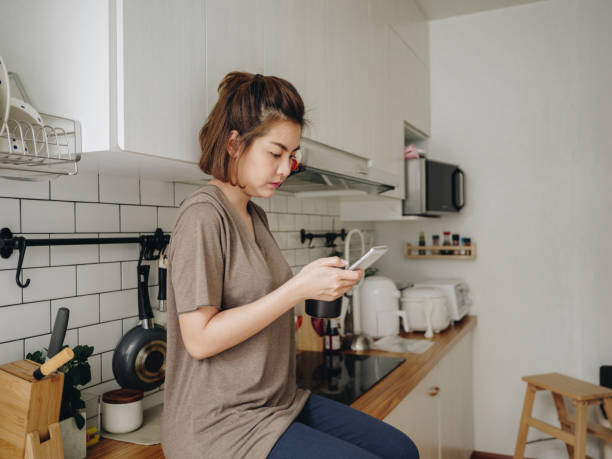 istock Teenage asian woman drinking coffee while sitting on kitchen counter and working on smart phone in morning at home. 1212332521
