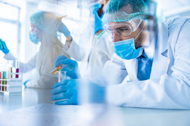 Male scientist working on scientific research in laboratory. Concentrated mid adult chemist working with poisonous liquid in laboratory. His colleagues are in the background. science research stock pictures, royalty-free photos & images
