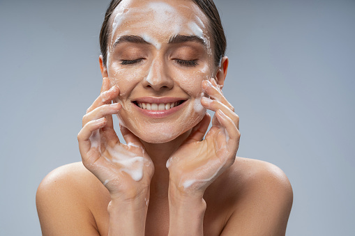Portrait of smiling attractive woman touching face by hands while cleansing the skin from makeup. Isolated on grey background. Cosmetics and care concept
