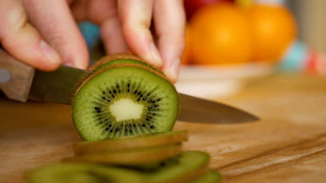 Man slicing kiwi with knife on wooden board with fruits and vegetables on background. Close-up.
