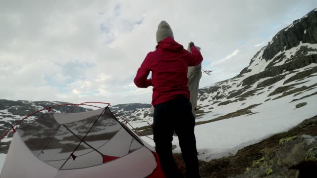 A young woman putting up the tent in the nearby of Trolltunga, Norway. Wild camping in the nature. Couple is having fun. They are surrounded by snow. Winter mountain climbing. Freedom and adventure.