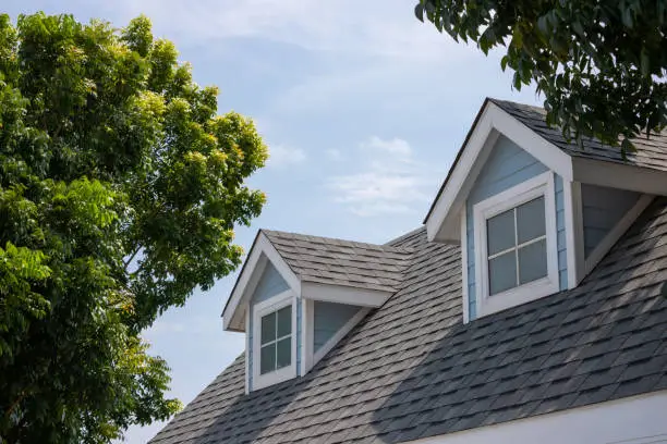 Photo of Roof shingles with garret house on top of the house among a lot of trees. dark asphalt tiles on the roof background.
