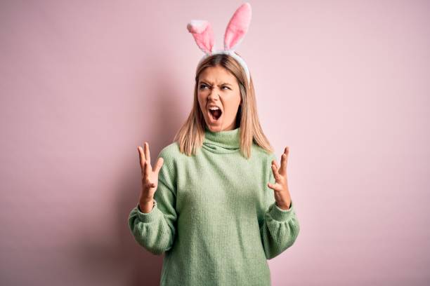 jeune belle femme utilisant des oreilles de lapin de pâques restant au-dessus de fond rose isolé fou et criant et criant avec l’expression agressive et les bras levés. concept de frustration. - easter rabbit bizarre women photos et images de collection