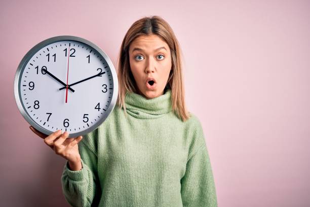 istock Young beautiful woman holding clock standing over isolated pink background scared in shock with a surprise face, afraid and excited with fear expression 1212376265