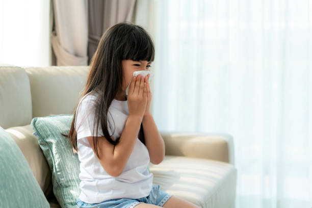 Asian child or kid girl sick and sad with sneezing on nose and cold cough on tissue paper because influenza and weak or virus bacteria from dust weather or smoke and kindergarten school for medical. stock photo