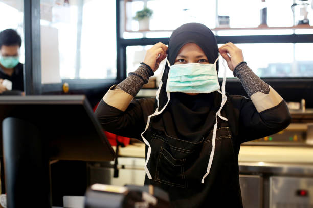 South East Asia: At The Airport A Muslim female adult is positioning and tying surgical mask strap at airport cafe in Malaysia. asian cashier stock pictures, royalty-free photos & images