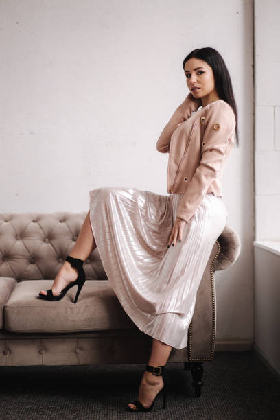 istock Beautiful brunette woman demonstrating spring clothes in studio. Model sitting on sofa. White background 1212396024