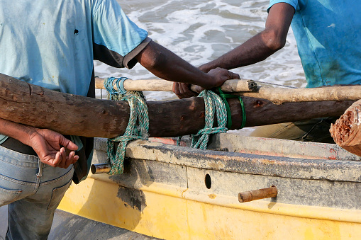 Two fishermen working together to pull their fishing boat out of the sea, after a fishing trip. They are leaning on a wooden pole that is tied onto the boat with green and white rope. They are standing facing towards the sea and pushing backwards.