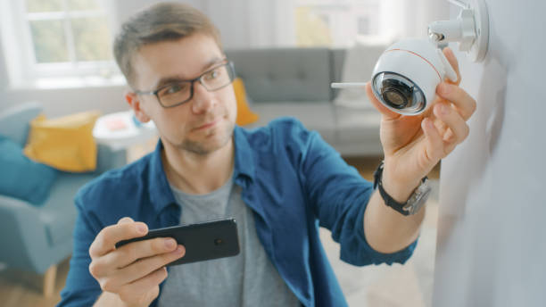 ung man i glasögon bär en blå skjorta justerar en modern wi-fi övervakningskamera med två antenner på en vit vägg hemma. han kollar videoflödet på sin smartphone. - security home bildbanksfoton och bilder