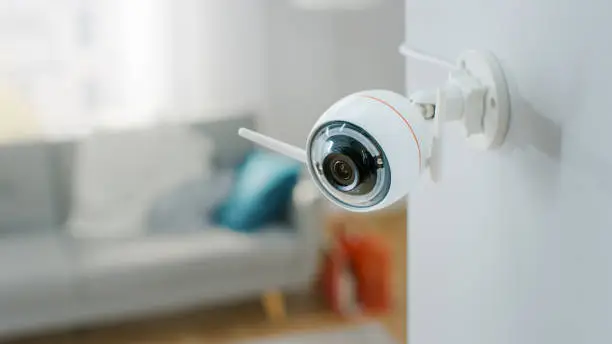 Close Up Object Shot of a Modern Wi-Fi Surveillance Camera with Two Antennas on a White Wall in a Cozy Apartment. Man is Sitting on a Sofa in the Background.