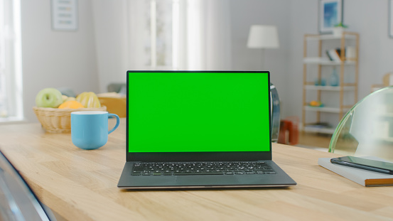 Shot of a Modern Laptop with a Horizontal Green Screen Mock Up on a Wooden Table at Home. Smartphone Lies on a Table Next to the Computer.