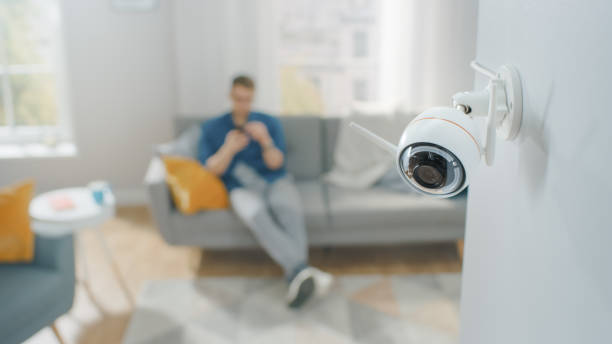 close up object shot de una moderna cámara de vigilancia wi-fi con dos antenas en una pared blanca en un apartamento acogedor. el hombre está sentado en un sofá en el fondo. - secrecy surveillance security system order fotografías e imágenes de stock