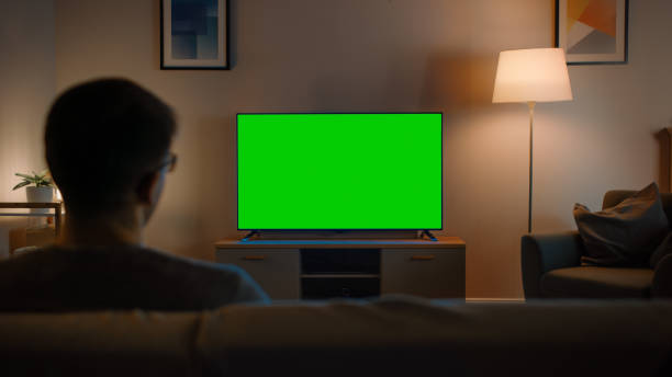 istock Young Man in Glasses is Sitting on a Sofa and Watching TV with Horizontal Green Screen Mock Up. It's Evening and Room at Home Has Working Lamps. 1212406193