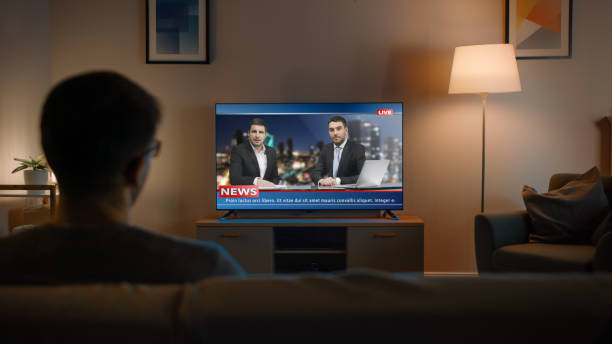 young man in glasses sitzt auf einem sofa und schaut mit live news fern. it es evening and room at home has working lamps. - nachrichtenereignis stock-fotos und bilder