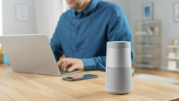 istock Close Up Shot of a Modern Silver Bluetooth Speaker Standing on a Table at Home. Man in the Background Sits at the Table and Works on His Laptop. Smartphone Lies on a Table Next to Him. 1212406241