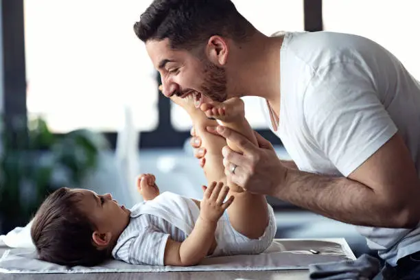 Photo of Smiling young father has fun with little baby while changing his nappy at home.