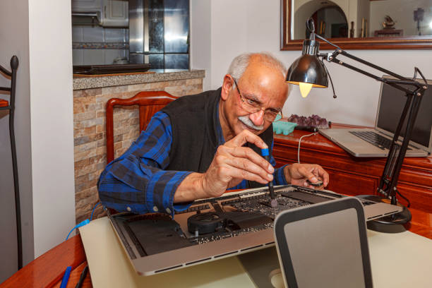 A 72 year old Asian Indian works at home to upgrade his personal computer. He has opened the computer and has just dismounted the Hard Drive. He is next working on dismantling the Power Supply module. He has a laptop computer open by his side, in case he needs to check any process on the Internet. There is also an electric table lamp on the table to light up the area where the work is being done. He is working on a rosewood dining table inside his home. He is casually dressed in a blue checked shirt with long sleeves. Horizontal format. Note to inspector: all logos and barcodes have been removed. un indio asiático de 72 años visto trabajando en una computadora abierta en casa para hacer una actualización; después de haber quitado el disco duro, ahora está trabajando en el desmantelamiento del módulo de fuente de alimentación - logos y códig