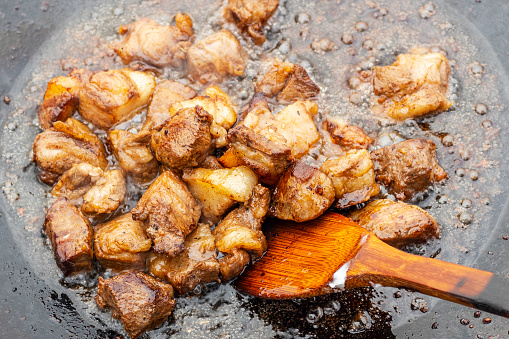 Pieces of red meat are fried in an old frying pan. Close up photo of meat, greaves, wooden kitchen spatula and splashes of fat.