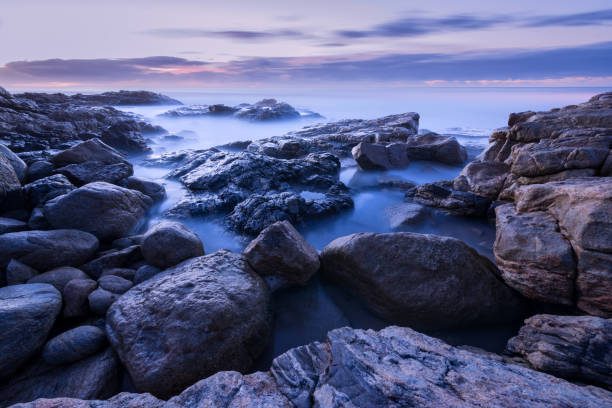 A pre-dawn seascape photograph of misty waves crashing on the rocks A pre-dawn seascape photograph of misty waves crashing on the rocks, with a purple, cloudy sky and rock formations in the foreground, taken by the South Coast in South Africa long shutter speed stock pictures, royalty-free photos & images