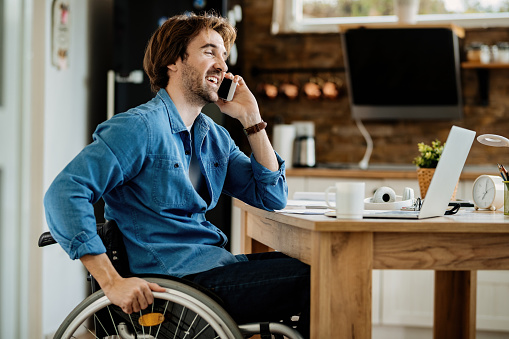 Young happy businessman in wheelchair working at home and talking on cell phone.