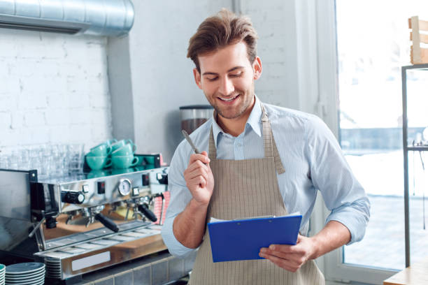 istock Young adult barista working in coffeeshop, making important notes in folder 1212441174