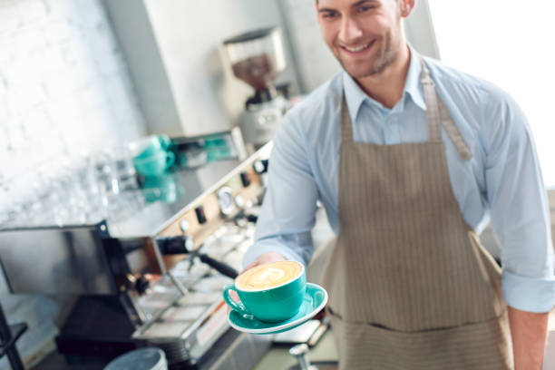 コーヒーショップで働く若い大人のバリスタ、コーヒーとカップを持っている - barista coffee coffee crop heart shape ストックフォトと画像