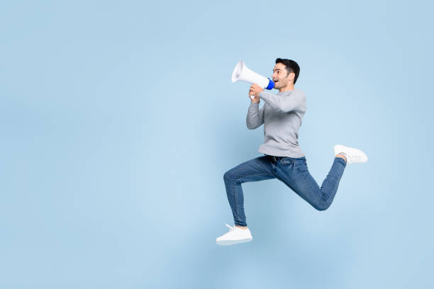 Young active man jumping and shouting on megaphone isolated on light blue background with copy space Young active man jumping and shouting on megaphone isolated on light blue background with copy space happy people audio stock pictures, royalty-free photos & images