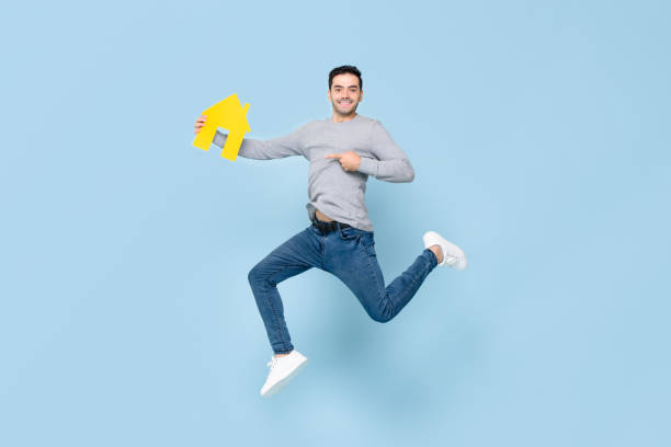 istock Happy handsome man jumping and pointing to yellow house model isolated on blue studio background 1212441248