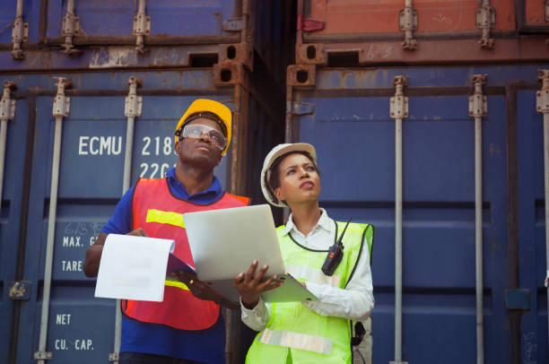 istock Worker man in hardhat and safety vest holding clipboard checklist and Female foreman using laptop control loading containers box from cargo 1212446979