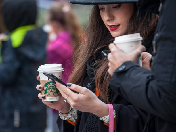 mujer joven, al estilo hipster, sosteniendo un teléfono inteligente mientras sostiene una taza de café con el logotipo de starbucks, una de las principales cadenas de café en el mundo. - starbucks coffee drink coffee cup fotografías e imágenes de stock
