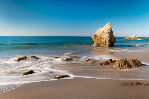 Looking down the shore of Malibu's iconic El Matador State Beach. It's a bright blue, cloudless, day as the waves wash up the sandy shore at the base of the cliffs and around the various boulders and rock formations. A long exposure makes the waves appear soft and flow-y. There are no clouds in the sky, and no people on the beach.