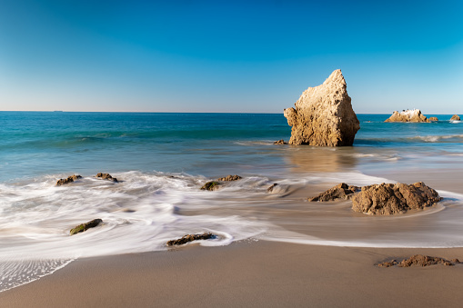 Looking down the shore of Malibu's iconic El Matador State Beach. It's a bright blue, cloudless, day as the waves wash up the sandy shore at the base of the cliffs and around the various boulders and rock formations. A long exposure makes the waves appear soft and flow-y. There are no clouds in the sky, and no people on the beach.