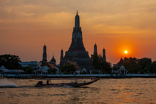 Longtail boat, with tourists, passing along the Chao Phraya River at sunset with the world-famous temple of Wat Arun silhouetted behind on the skyline in Bangkok. Wat Arun has a spectacular high decorative stupa set in the heart of the temple complex as well as being in the heart of the city.