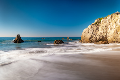 Looking down the shore of Malibu's El Matador Beach. The Malibu cliffs descend into the waves and a few boulders rise out of the water. A long exposure makes the waves and foam feel soft and motion blurred as the splash on the rocks. The sky is rich vibrant blue, the water has a turquoise tint, and the rocks are warm and bright. There are no clouds in the sky and no people on the beach.