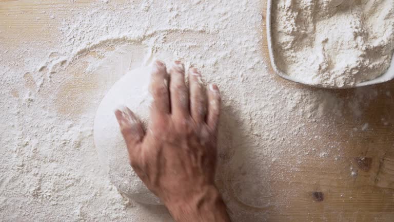 Slow motion top view of man's hands sprinkling flour over pizza dough and massaging it to prepare it for leavening at home on a light wooden board with white flour