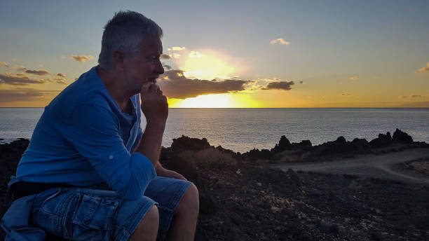 istock Side view of a smiling elderly man sitting along the cliff while looking pensively at the horizon on the water. The rays of the setting sun. Freedom and relaxation concept. 1212501200