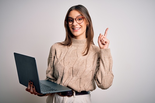Young beautiful brunette woman working using laptop over isolated white background surprised with an idea or question pointing finger with happy face, number one