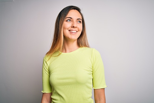 Beautiful blonde woman with blue eyes wearing green casual t-shirt over white background looking away to side with smile on face, natural expression. Laughing confident.